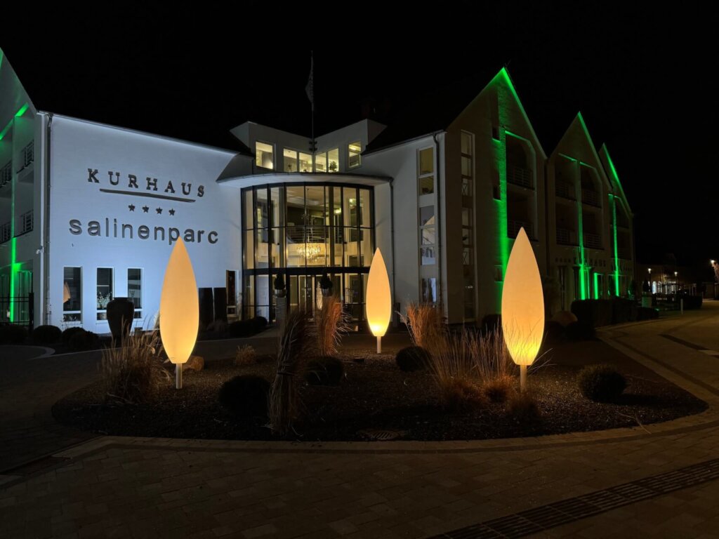 Ein beleuchtetes Kurhaus mit der Aufschrift „salinenparc“ in der Nacht. Vor dem Gebäude stehen moderne, tropfenförmige Lichtskulpturen, während die Fassade in Grün- und Weißtönen angestrahlt wird in Bad Westernkotten