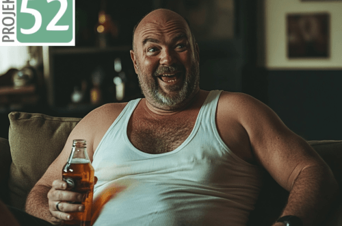 Projekt 52- Veränderung: Mein Ruhestand. Ein fröhlicher, bärtiger Mann mit Glatze sitzt entspannt auf einem Sofa, trägt ein weißes Unterhemd und hält eine Flasche Bier in der Hand. Er lacht herzlich und wirkt ausgelassen. Der Hintergrund zeigt ein gemütlich eingerichtetes Wohnzimmer mit Regalen und Dekoration. Mein Ruhestand.