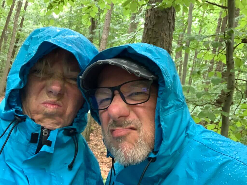 Ein Mann und eine Frau stehen dicht beieinander, beide tragen blaue Regenjacken mit hochgezogenen Kapuzen. Ihre Gesichter zeigen grimmige, missmutige Ausdrücke, als wären sie vom Wetter genervt. Der Mann trägt eine Brille und eine dunkle Baseballkappe unter der Kapuze. Auf den Jacken sind Wassertropfen zu sehen, was darauf hindeutet, dass es regnet oder geregnet hat. Der Hintergrund wurde entfernt, sodass nur die Personen im Fokus stehen.