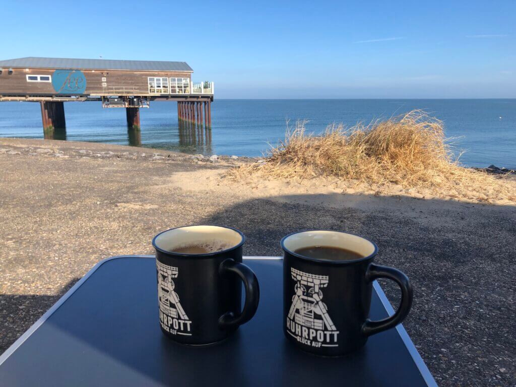 Zwei schwarze Kaffeetassen mit „Ruhrpott Glück Auf“-Aufdruck stehen auf einem Campingtisch mit Blick auf das Meer. Im Hintergrund ist ein auf Stelzen stehendes Holzgebäude mit der Aufschrift „Fee“ zu sehen. Der Himmel ist blau, das Meer ruhig, und neben dem Tisch wachsen Sanddünen mit trockenem Gras.
