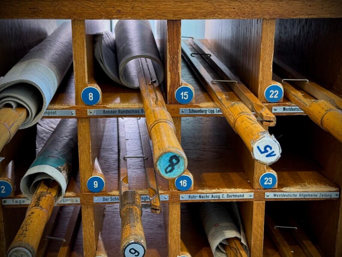 Wochenendtrip nach Bad Salzuflen und die VitaSol-Therme. Eine historische Zeitungsablage aus Holz mit nummerierten Fächern, in denen Zeitungen auf Holzstäben aufgerollt sind. Die Nahaufnahmen zeigen die Detailbeschriftung der Zeitungsrollen, während die Gesamtaufnahme die gesamte Schrankkonstruktion mit Glastüren und einem angrenzenden Bücherregal in einem modernen Leseraum darstellt.
