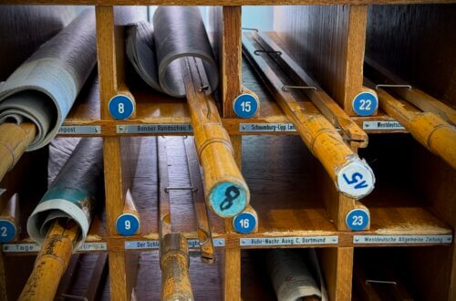 Wochenendtrip nach Bad Salzuflen und die VitaSol-Therme. Eine historische Zeitungsablage aus Holz mit nummerierten Fächern, in denen Zeitungen auf Holzstäben aufgerollt sind. Die Nahaufnahmen zeigen die Detailbeschriftung der Zeitungsrollen, während die Gesamtaufnahme die gesamte Schrankkonstruktion mit Glastüren und einem angrenzenden Bücherregal in einem modernen Leseraum darstellt.