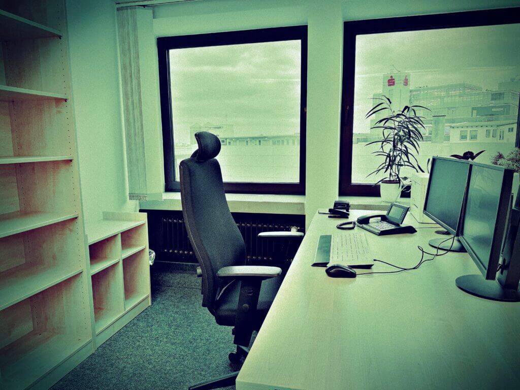 Modernes Büro mit leerem Schreibtisch, ergonomischem Bürostuhl und zwei Monitoren vor großen Fenstern mit Blick auf eine Stadtlandschaft. Links steht ein leeres Regal, eine Zimmerpflanze bringt etwas Grün ins Büro. Eintritt in den Ruhestand.