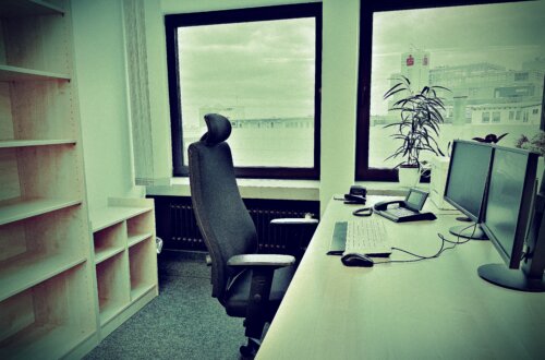 Modernes Büro mit leerem Schreibtisch, ergonomischem Bürostuhl und zwei Monitoren vor großen Fenstern mit Blick auf eine Stadtlandschaft. Links steht ein leeres Regal, eine Zimmerpflanze bringt etwas Grün ins Büro. Eintritt in den Ruhestand.
