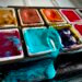 Kreativanfall - Nahaufnahme eines alten Aquarellkastens mit intensiven Farben – auslaufendes Türkis sorgt für kreative Unordnung – Symbol für künstlerische Inspiration, Malfreude und analoge Kreativität im Ruhestand.