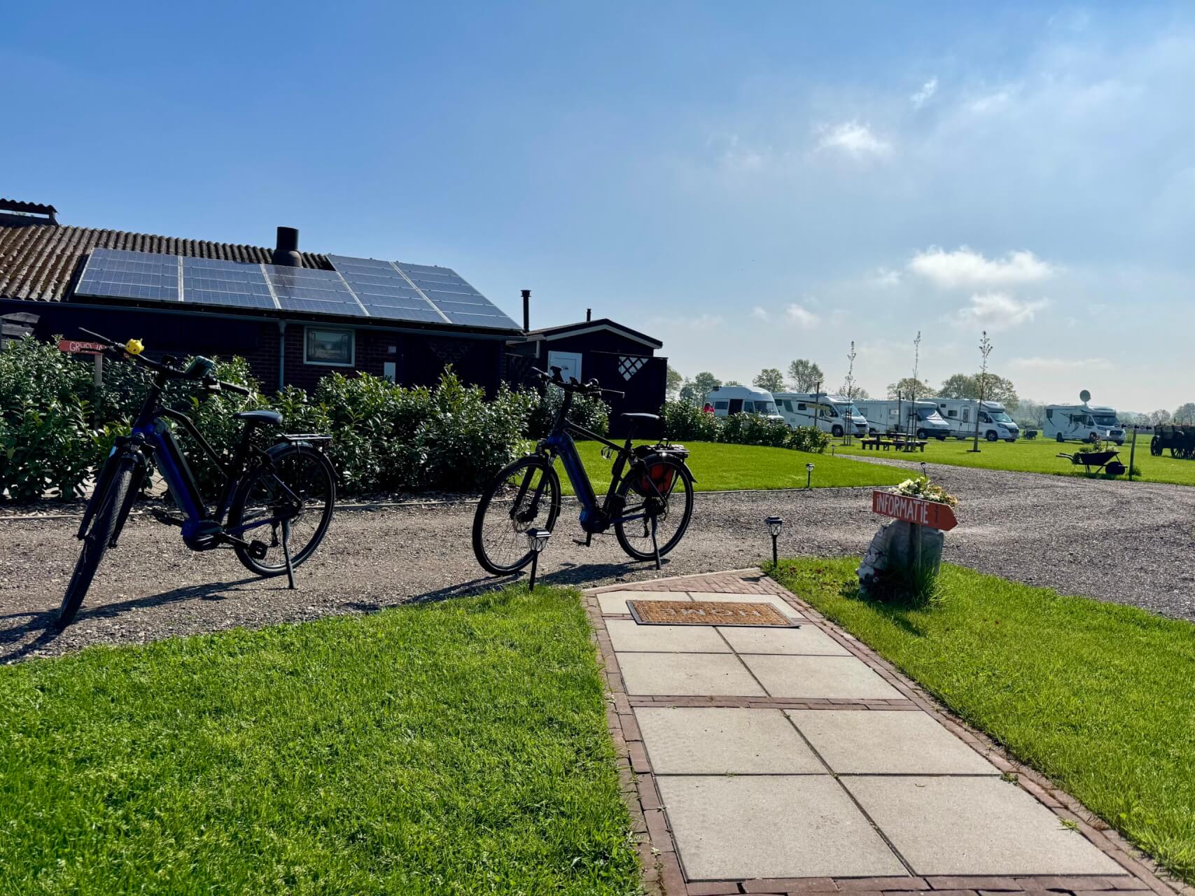 Wohnmobil auf einem ruhigen Campingplatz mit aufgebautem Campingtisch und Stühlen, umgeben von blühenden Pflanzen unter leicht bewölktem Himmel. Im Vordergrund stehen zwei Fahrräder