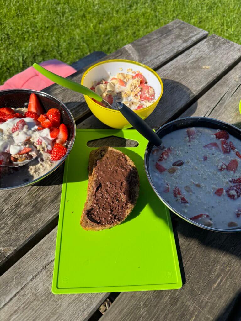 Draußen auf einem Holztisch stehen drei Schüsseln mit Müsli, Joghurt und frischen Erdbeeren. In der Mitte liegt auf einem grünen Schneidebrett eine Scheibe Brot mit Schokoladenaufstrich. Die Szene ist sonnig, im Hintergrund ist grünes Gras zu sehen. Ein Messer liegt auf einer der Schüsseln. Es wirkt wie ein gemütliches Frühstück im Freien.
