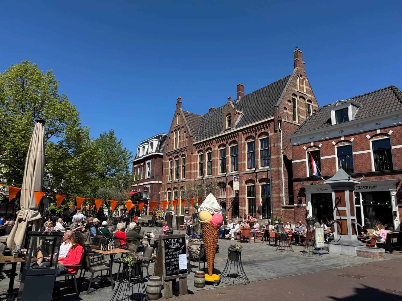 Belebter Platz mit Straßencafés in einer historischen Altstadt bei sonnigem Wetter. Menschen sitzen an Tischen unter freiem Himmel, orangefarbene Wimpel sind gespannt. Im Vordergrund eine große Eistüte als Werbefigur. Im Hintergrund stehen Backsteingebäude im niederländischen Stil mit Treppengiebeln und niederländischer Flagge.