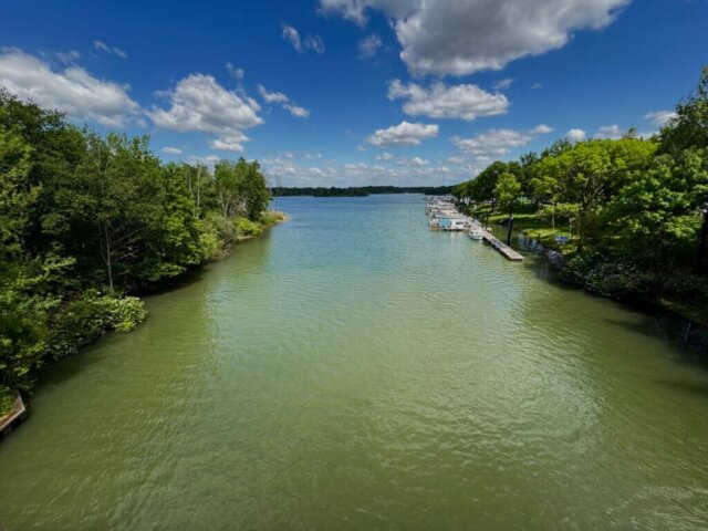 Blick auf das grüne Flussufer mit gepflegten Wiesen, Bäumen und einzelnen Bootsstegen an einem breiten Fluss. Im Vordergrund fährt ein Jet-Ski über das Wasser und hinterlässt eine weiße Spur. Der Himmel ist mit vielen Wolken bedeckt, dazwischen zeigen sich blaue Stellen. Im Hintergrund sind Felder und eine Bahntrasse zu sehen. Leukermeer, Radtour Kevelaer