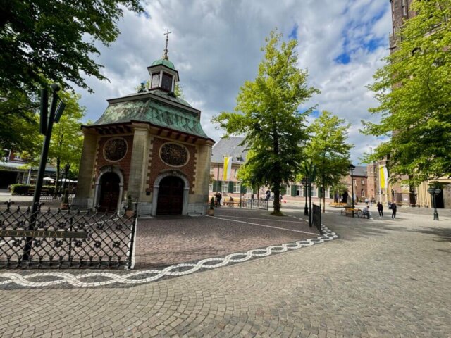 Historischer Platz mit Kopfsteinpflaster und einer kleinen achteckigen Kapelle mit grünem Kupferdach und kunstvollen Wappenreliefs in der Altstadt von Kevelaer. Umgeben von Bäumen, Sitzbänken und gelben Fahnen, während im Hintergrund Menschen über den Platz gehen. Der Himmel ist teilweise bewölkt mit Licht- und Schattenwechsel. Radtour Kevelaer.