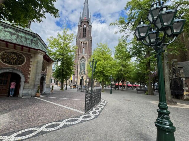 Kopfsteinplatz in Kevelaer mit Blick auf die neugotische Marienbasilika und die achteckige Gnadenkapelle im Vordergrund. Eine Frau betritt die Kapelle, daneben eine kunstvolle Laterne und gepflasterte Wege mit weißem Mosaikmuster. Der Platz ist von Bäumen gesäumt, im Hintergrund sind Straßencafés zu sehen, teils unter bewölktem Himmel. Radtour Keveler