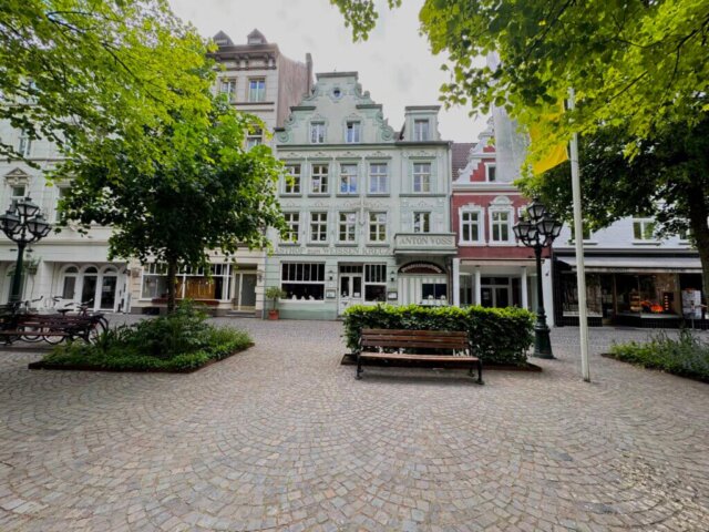 Historische Häuserfassaden in der Innenstadt von Kevelaer mit dem Gasthof „Zum weißen Kreuz“ im Zentrum. Vor dem Gebäude befindet sich ein kleiner, gepflasterter Platz mit Sitzbänken, Laternen und grüner Bepflanzung. Die Szene ist von Bäumen eingerahmt, im Hintergrund erkennt man weitere Geschäfte und Cafés. Radtour Kevelaer