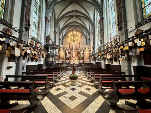 Blick ins Innere der Kerzenkapelle St. Michael in Kevelaer mit barock gestaltetem Altarraum, hohen Spitzbogenfenstern mit buntem Glas und dunklen Holzbänken. Die Seitenwände sind reich geschmückt mit Pilgerabzeichen, Wappen und Votivtafeln. Der Boden ist mit schwarz-weißem Marmor-Muster ausgelegt, eine Osterkerze steht im Mittelgang. Radtour Kevelaer