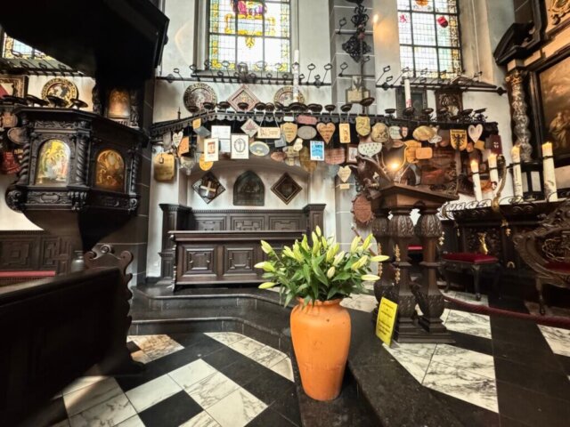 Detailansicht des Chorraums in der Kerzenkapelle St. Michael Kevelaer mit einem kunstvoll geschnitzten Holzkanzel, reich verzierten Wänden voller Votiv- und Pilgertafeln, sowie einem Rednerpult mit geschnitztem Adler. Im Vordergrund steht eine große Tonvase mit weißen Lilien auf dem schwarz-weiß gemusterten Marmorboden. Farbige Bleiglasfenster lassen Licht herein