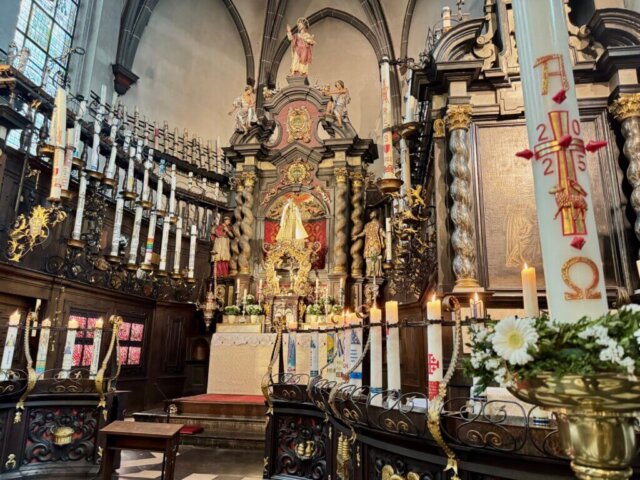 Detailansicht des Chorraums in der Kerzenkapelle Kevelaer mit einem kunstvoll geschnitzten Holzkanzel, reich verzierten Wänden voller Votiv- und Pilgertafeln, sowie einem Rednerpult mit geschnitztem Adler. Im Vordergrund steht eine große Tonvase mit weißen Lilien auf dem schwarz-weiß gemusterten Marmorboden. Farbige Bleiglasfenster lassen Licht herein