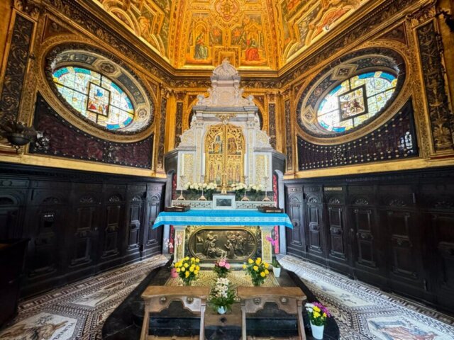 Blick auf den reich verzierten Altarraum in der Sakramentskapelle der Gnadenkapelle Kevelaer. Der Raum ist mit goldenen Wandmalereien, Mosaikfußboden und zwei großen ovalen Buntglasfenstern geschmückt. Vor dem Altar stehen Holzbänke und Blumenschmuck mit gelben und rosa Blumen, die eine Atmosphäre der Andacht und Ruhe erzeugen.