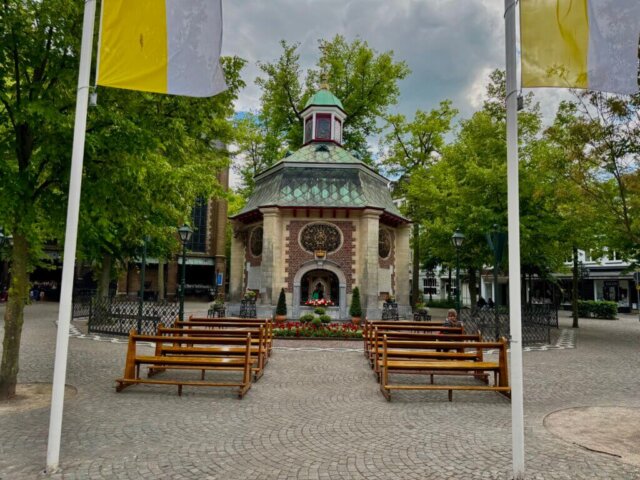 Frontalansicht der Gnadenkapelle in Kevelaer mit grünem Kupferdach und achteckiger Architektur, umgeben von Bäumen und gelb-weißen Fahnen. Vor der Kapelle stehen mehrere hölzerne Bänke, auf denen einzelne Menschen sitzen und beten. Der Platz ist gepflastert und mit Blumenbeeten geschmückt, der Himmel ist leicht bewölkt.