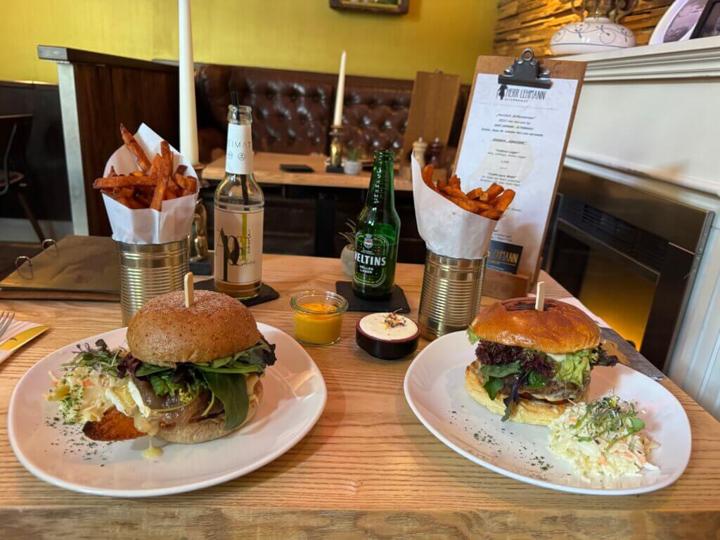 Zwei liebevoll angerichtete Burger mit Süßkartoffelpommes, Dips und Cole Slaw auf weißen Tellern in einem stilvollen Restaurantambiente. Auf dem Tisch stehen außerdem eine Veltins-Bierflasche, eine Bio-Limonade und eine Speisekarte auf einem Klemmbrett. Die Burger sind mit frischem Salat, Zwiebeln und hausgemachten Brötchen serviert.