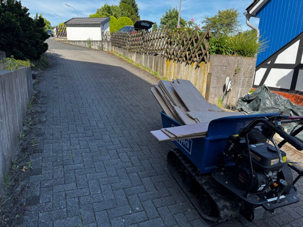 Scheppach-Kettendumper mit Holzplatten beladen fährt eine steile gepflasterte Auffahrt hinauf entlang eines Fachwerkhauses mit blauem Anstrich bei sonnigem Wetter.