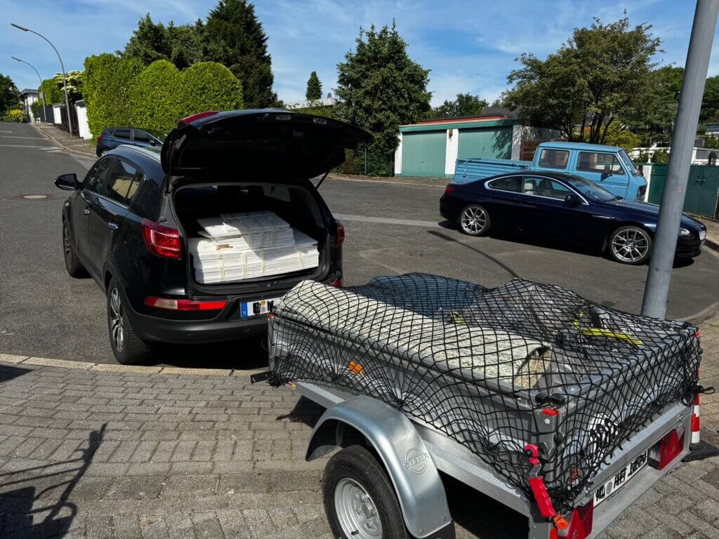 Schwarzer SUV mit offenem Kofferraum, voll beladen mit weißen Wandfliesen, daneben ein Anhänger mit gesicherter Ladung auf einem ruhigen Wohnstraßenparkplatz bei sonnigem Wetter.