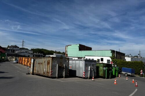 Recyclinghof mit verschiedenen Containern für Altmetall, Sperrmüll und Wertstoffe auf einem asphaltierten Platz bei Industriegebäuden und unter blauem Himmel.