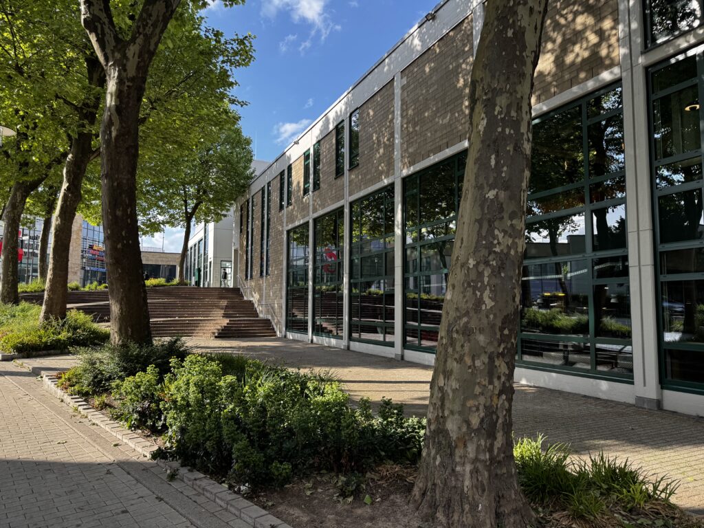 Forum Velbert mit großer Fensterfront und heller Klinkerfassade, umgeben von schattenspendenden Bäumen und gepflegten Grünflächen an einem sonnigen Tag mit blauem Himmel. Poetry Slam Velbert