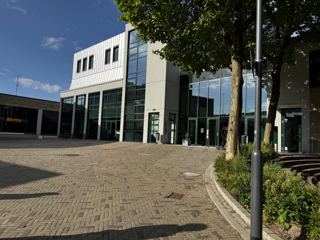 Moderne Glasfassade des Forum Velbert mit barrierefreiem Eingangsbereich, umliegenden Bäumen und großzügigem gepflasterten Vorplatz bei sonnigem Wetter; im Hintergrund ein Netto-Supermarkt. Poetry Slam Velbert