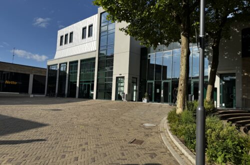 Moderne Glasfassade des Forum Velbert mit barrierefreiem Eingangsbereich, umliegenden Bäumen und großzügigem gepflasterten Vorplatz bei sonnigem Wetter; im Hintergrund ein Netto-Supermarkt. Poetry Slam Velbert