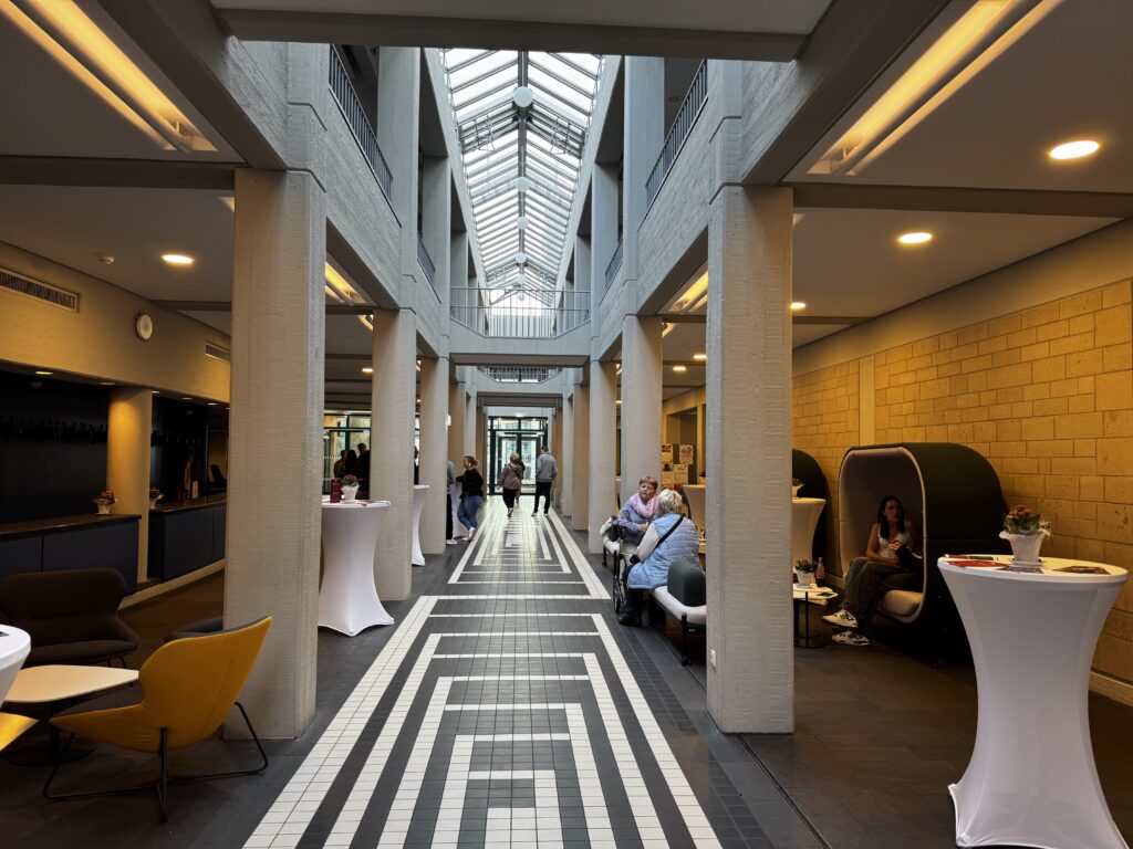 Innenansicht des Forum Velbert mit hoher Glasdecke, Sitzgelegenheiten entlang eines schwarz-weiß gemusterten Flurs, Stehtischen mit weißen Hussen und Personen im Gespräch oder beim Entspannen. Poetry Slam Velbert