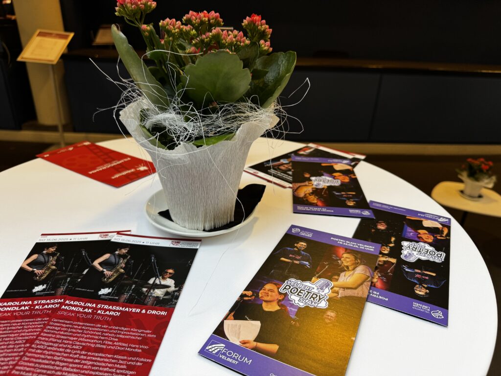 Runder Stehtisch mit Dekoblume und Veranstaltungsflyern für ein Jazzkonzert und einen Poetry Slam im Forum Velbert, aufgenommen in einem modernen Foyer mit stilvoller Atmosphäre.