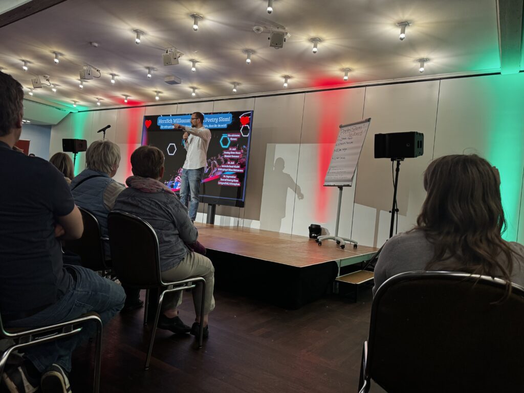 Poetry Slam Veranstaltung im Forum Velbert mit einem Moderator auf einer beleuchteten Bühne vor Publikum, großer Bildschirm mit Veranstaltungsinfos, Flipchart und stimmungsvollem Licht in Rot und Grün.