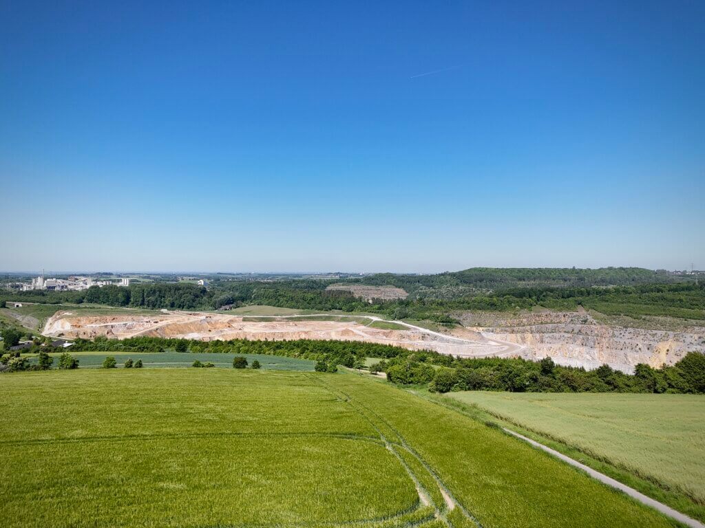 Panoramaaufnahme einer offenen Landschaft mit einem großen Kalksteinbruch in der Bildmitte. Im Vordergrund sind grüne Felder und ein schmaler Weg zu sehen. Im Hintergrund erstreckt sich eine industrielle Anlage links sowie bewaldete Hügel und ein klarer blauer Himmel ohne Wolken.