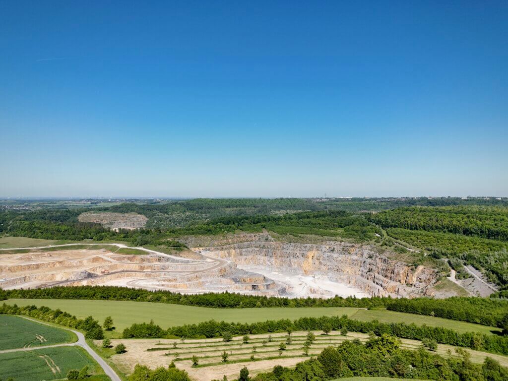 Panoramaaufnahme einer offenen Landschaft mit einem großen Kalksteinbruch in der Bildmitte. Im Vordergrund sind grüne Felder und ein schmaler Weg zu sehen. Im Hintergrund erstreckt sich eine industrielle Anlage links sowie bewaldete Hügel und ein klarer blauer Himmel ohne Wolken.