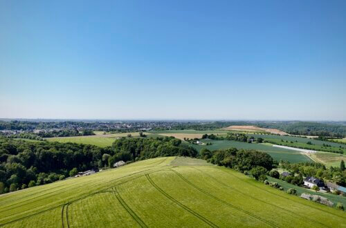 Luftaufnahme einer sanft hügeligen Landschaft mit grünen Feldern im Vordergrund. Im Mittelgrund verlaufen Baumreihen und vereinzelte Häuser. In der Ferne liegt eine weitläufige Ortschaft, umgeben von weiteren Feldern und Wäldern. Darüber spannt sich ein wolkenloser, blauer Himmel.