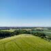 Luftaufnahme einer sanft hügeligen Landschaft mit grünen Feldern im Vordergrund. Im Mittelgrund verlaufen Baumreihen und vereinzelte Häuser. In der Ferne liegt eine weitläufige Ortschaft, umgeben von weiteren Feldern und Wäldern. Darüber spannt sich ein wolkenloser, blauer Himmel.