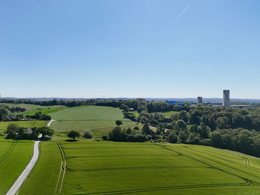 Kontraste auf dem Land: Zwischen sattgrünen Feldern und sanften Hügeln ragen Industrieanlagen in den Himmel. Ein schmaler Weg führt durch die idyllische Landschaft und verbindet Natur mit Zivilisation – ein Bild des modernen ländlichen Raum