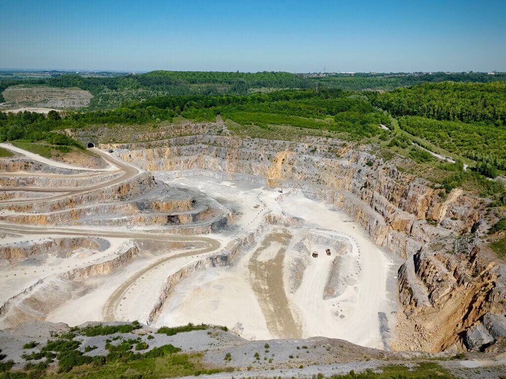 Luftaufnahme eines großen Kalksteinbruchs mit terrassenförmig angelegten Abbauflächen. In der Mitte des Steinbruchs sind helle, sandige Wege und zwei Baufahrzeuge zu erkennen. Der Steinbruch ist von dichter, grüner Waldlandschaft umgeben, darüber ein klarer blauer Himmel.