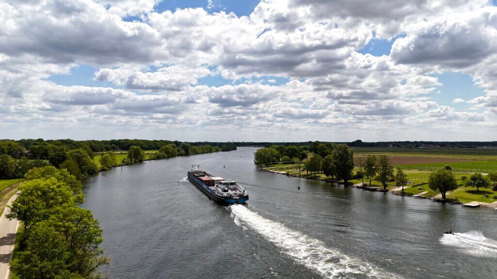Ein Frachtschiff fährt auf einem breiten Fluss durch eine grüne Landschaft mit Bäumen und Feldern unter einem teils bewölkten Himmel. Am Ufer sind Spazierwege, Liegewiesen und ein kleiner Hafenbereich zu sehen. Das Schiff hinterlässt eine weiße Heckwelle im Wasser. Radtour Kevelaer