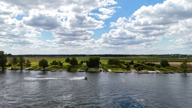 Blick auf das grüne Flussufer mit gepflegten Wiesen, Bäumen und einzelnen Bootsstegen an einem breiten Fluss. Im Vordergrund fährt ein Jet-Ski über das Wasser und hinterlässt eine weiße Spur. Der Himmel ist mit vielen Wolken bedeckt, dazwischen zeigen sich blaue Stellen. Im Hintergrund sind Felder und eine Bahntrasse zu sehen. Radtour Kevelaer