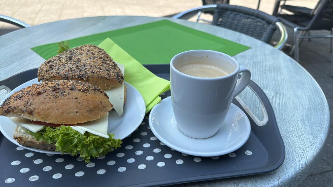 Frühstück im Café: Zwei Vollkornbrötchen mit Käse, Tomate und Salat auf einem weißen Teller neben einer Tasse Kaffee mit Crema, serviert auf einem Tablett im Außenbereich eines Cafés mit grünem Platzset auf dem Tisch. Rentnerleben kann ich