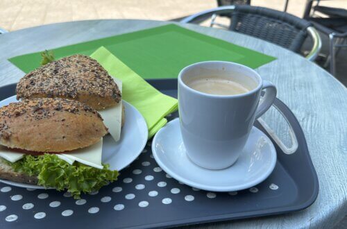 Frühstück im Café: Zwei Vollkornbrötchen mit Käse, Tomate und Salat auf einem weißen Teller neben einer Tasse Kaffee mit Crema, serviert auf einem Tablett im Außenbereich eines Cafés mit grünem Platzset auf dem Tisch. Rentnerleben kann ich