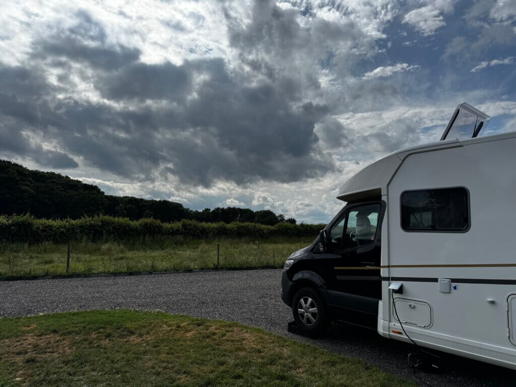 Modernes Wohnmobil auf einem Campingplatz in Beesel, Niederlande, bei sonnigem Wetter am 26. Juni. Temperatur 23 °C, Sonnenaufgang um 5:21 Uhr, Sonnenuntergang um 21:55 Uhr. Der Mercedes-Camper steht auf einer Parzelle mit Markise, Campingstühlen und Tisch, umgeben von grünen Bäumen.“