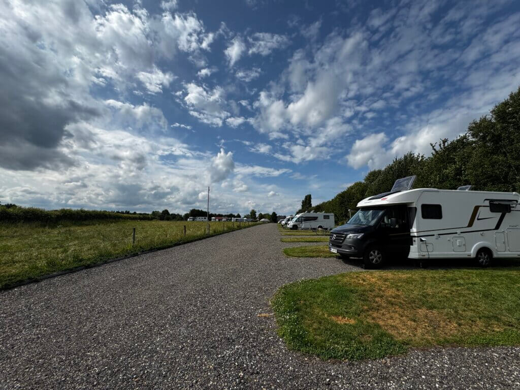 Modernes Wohnmobil auf einem Campingplatz in Beesel, Niederlande, bei sonnigem Wetter am 26. Juni. Temperatur 23 °C, Sonnenaufgang um 5:21 Uhr, Sonnenuntergang um 21:55 Uhr. Der Mercedes-Camper steht auf einer Parzelle mit Markise, Campingstühlen und Tisch, umgeben von grünen Bäumen.“
