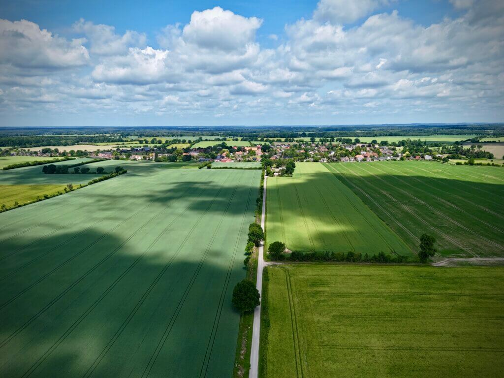 Ländliche Dorfansicht in Norddeutschland – Drohnenaufnahme zeigt eine schmale Landstraße, die durch weite grüne Getreidefelder zu einem idyllisch gelegenen Dorf mit roten Ziegeldächern führt, unter einem Himmel mit Sonne und Wolken.
