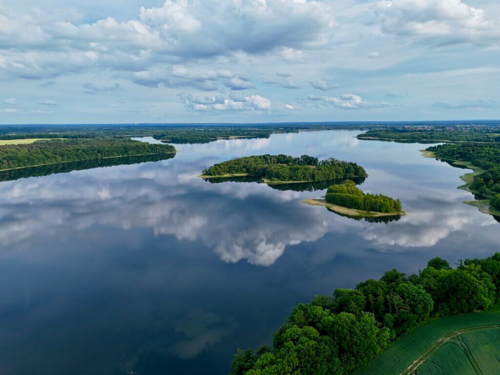 Drohnenaufnahme eines großen Sees mit bewaldeten Inseln in Norddeutschland – das ruhige Wasser spiegelt die Wolken am Himmel, umgeben von dichter Natur und sanften Uferlinien, mit weiter Sicht über die grüne Landschaft bis zum Horizont. Schaalsee