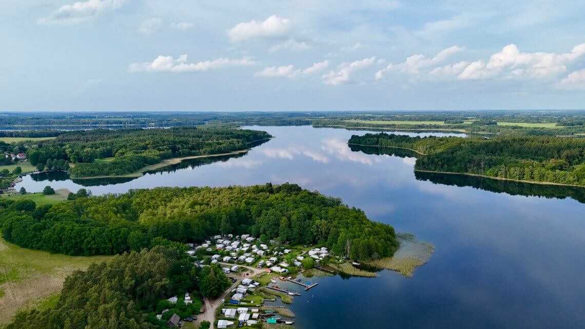 Drohnenaufnahme eines idyllischen Campingplatzes am Seeufer in Norddeutschland – zahlreiche Wohnwagen und Zelte liegen eingebettet zwischen Bäumen direkt am klaren Wasser, das von bewaldeten Ufern und kleinen Halbinseln umgeben ist, mit weitreichender Aussicht auf die Natur und den Himmel mit leichten Wolken. Schaalsee