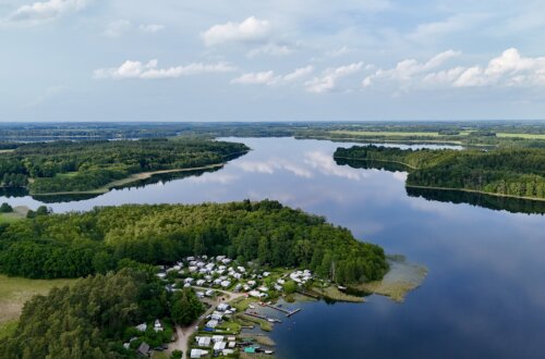 Drohnenaufnahme eines idyllischen Campingplatzes am Seeufer in Norddeutschland – zahlreiche Wohnwagen und Zelte liegen eingebettet zwischen Bäumen direkt am klaren Wasser, das von bewaldeten Ufern und kleinen Halbinseln umgeben ist, mit weitreichender Aussicht auf die Natur und den Himmel mit leichten Wolken. Schaalsee