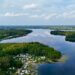 Drohnenaufnahme eines idyllischen Campingplatzes am Seeufer in Norddeutschland – zahlreiche Wohnwagen und Zelte liegen eingebettet zwischen Bäumen direkt am klaren Wasser, das von bewaldeten Ufern und kleinen Halbinseln umgeben ist, mit weitreichender Aussicht auf die Natur und den Himmel mit leichten Wolken. Schaalsee