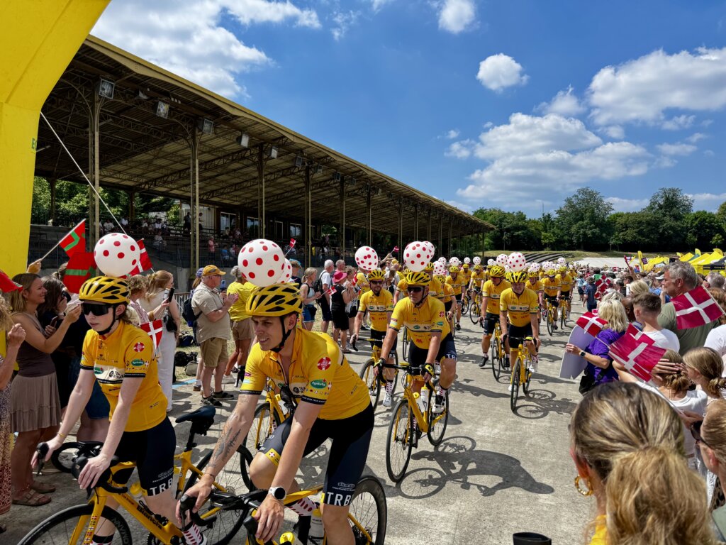 Große Gruppe von Radfahrenden in gelben Trikots, Helmen, Umhängen und Masken als Superhelden verkleidet, bei einer sportlichen Parade im Stadion – fröhliche Atmosphäre mit jubelnden Zuschauern am Rand, blauem Himmel und internationaler Beteiligung, inklusive dänischer Flaggen.