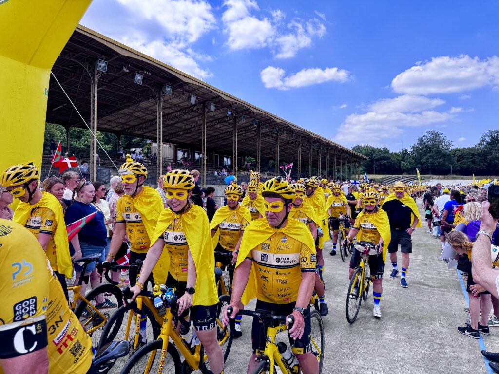 Große Gruppe von Radfahrenden in gelben Trikots, Helmen, Umhängen und Masken als Superhelden verkleidet, bei einer sportlichen Parade im Stadion – fröhliche Atmosphäre mit jubelnden Zuschauern am Rand, blauem Himmel und internationaler Beteiligung, inklusive dänischer Flaggen.
